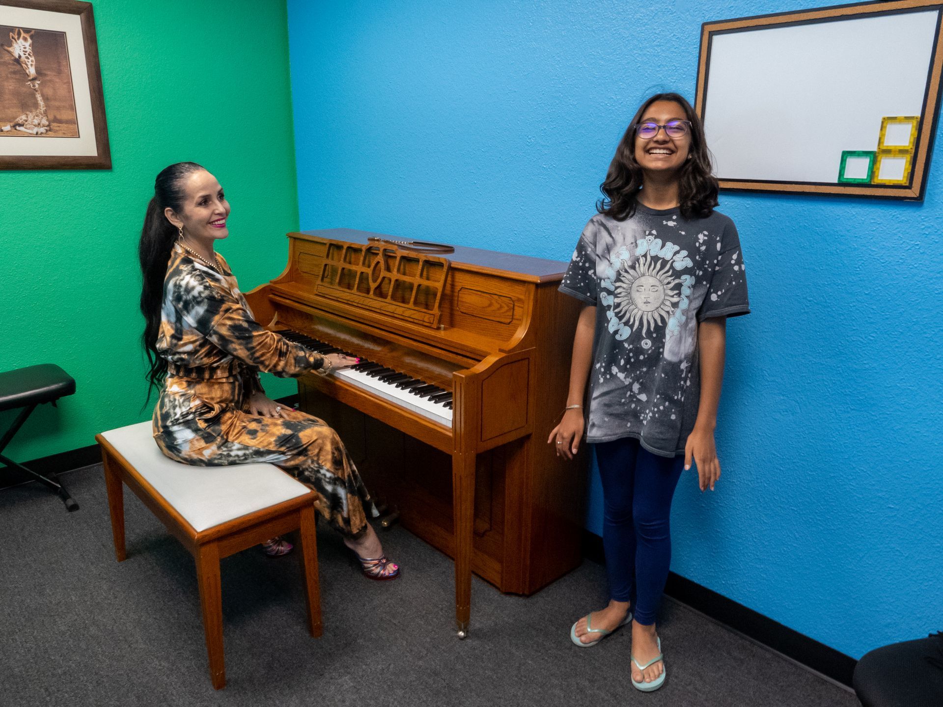 Two people smiling in a colorful room, one seated at an upright piano and one standing nearby.